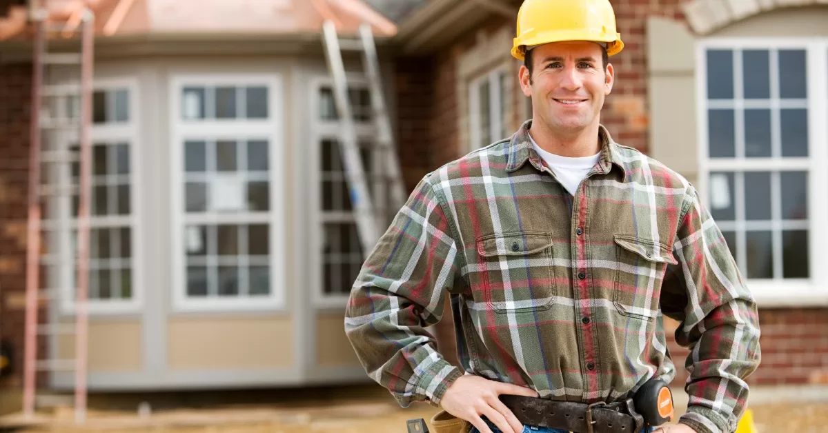 A man wearing a yellow hard hat and toolbelt stands in front of a home under renovation while smiling.