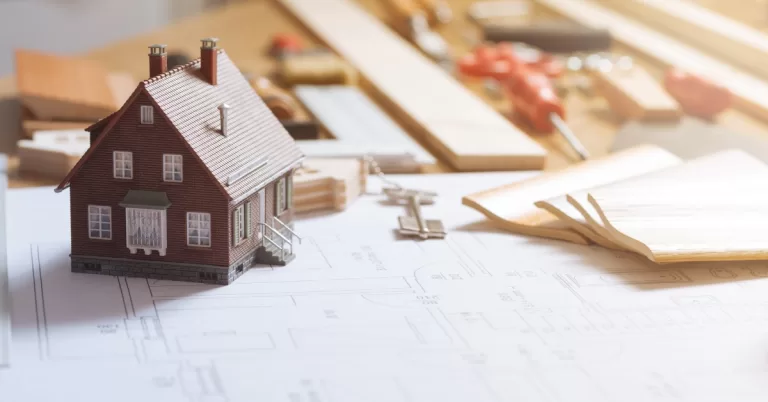 A small figure of a brick house sits on top of visual plans for a home renovation near supplies and tools.