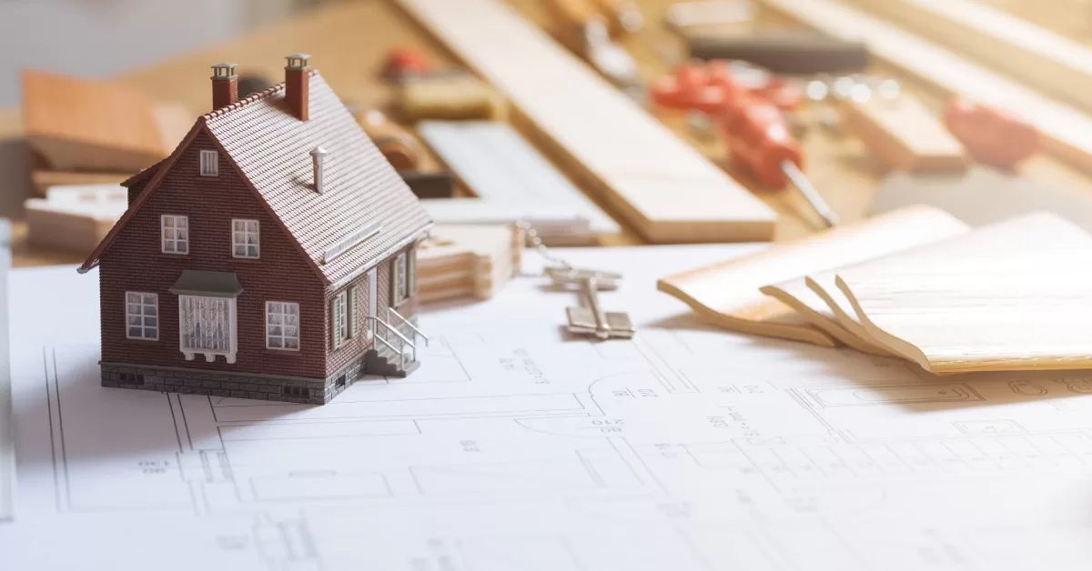 A small figure of a brick house sits on top of visual plans for a home renovation near supplies and tools.