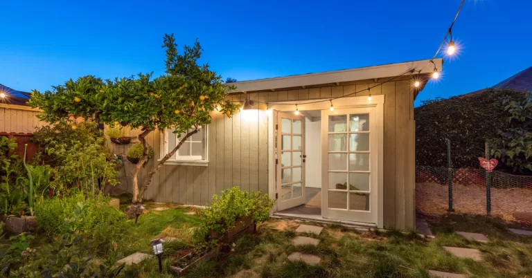 A small guest house with open French doors in a backyard at dusk, with lit string lights attached to the top overhang.