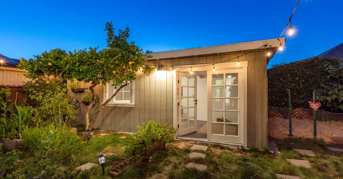 A small guest house with open French doors in a backyard at dusk, with lit string lights attached to the top overhang.
