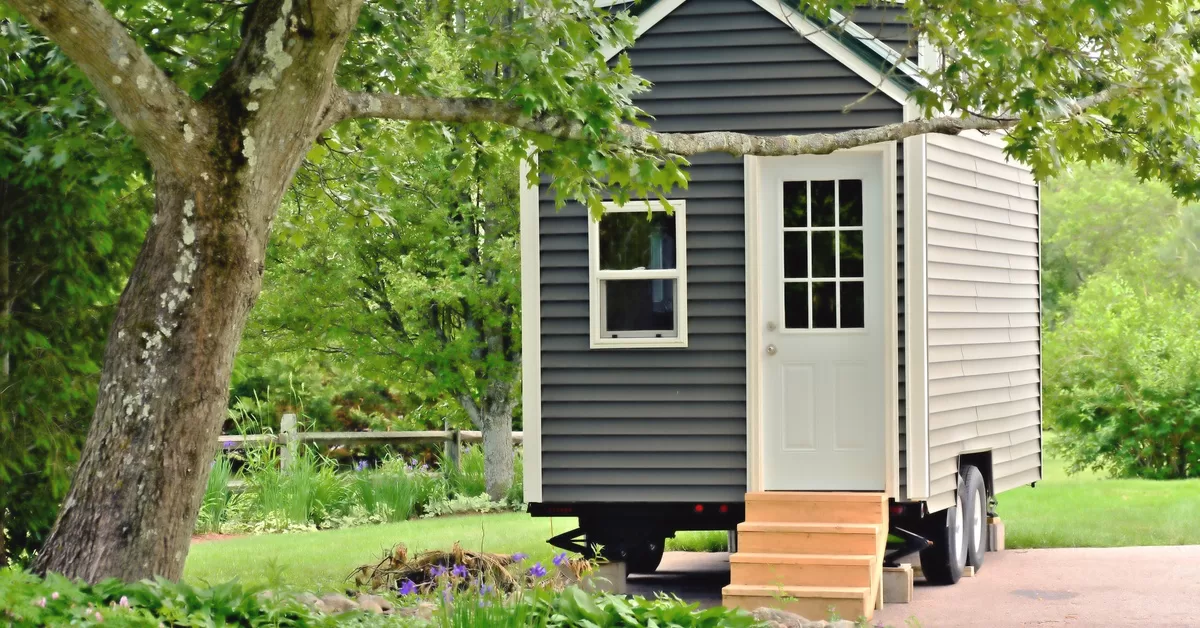 A tiny home with grey siding built on a trailer. Small steps lead to a white front door with a paned window.