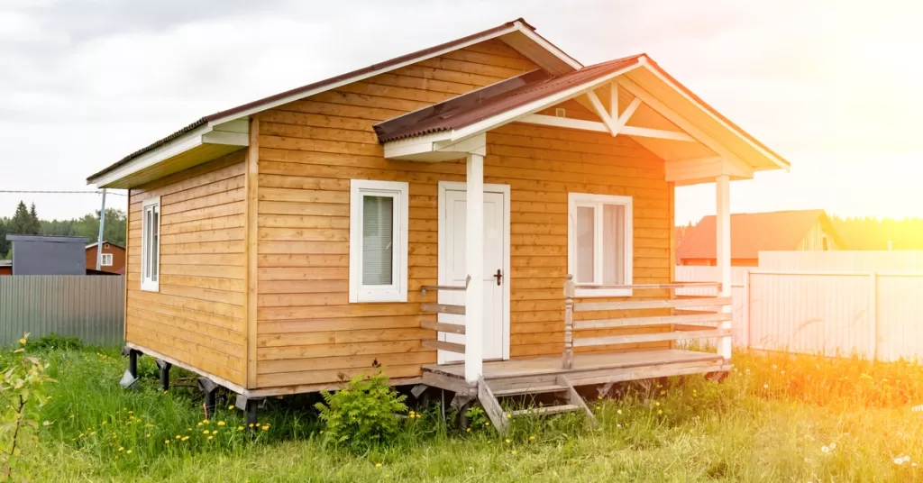 A tiny home with natural wood siding and a covered front porch in a grassy backyard with sunlight shining through.