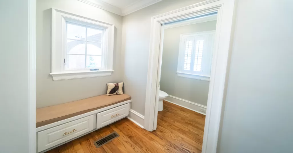 A mudroom features a padded bench and a dog pillow beneath a window, with a doorway leading to a half bathroom.