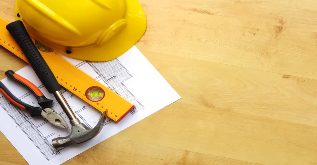 Tools, including a yellow construction helmet, blueprints, a level, a hammer, and pliers, lay out on a table.
