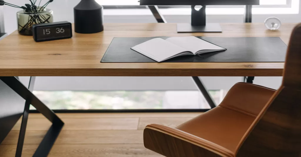A wooden desk with black legs, lit by sunlight. An open notebook sits on the desk, with a brown chair angled away.
