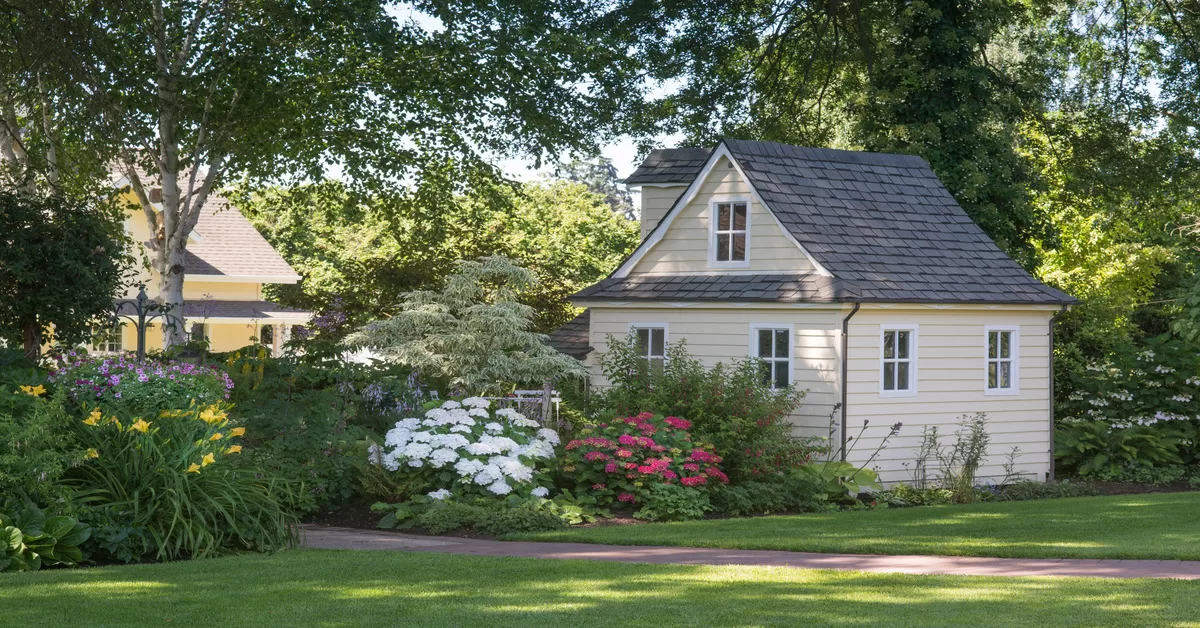 A small yellow building with gray shingles, featuring a red brick pathway, green grass, and surrounding foliage.