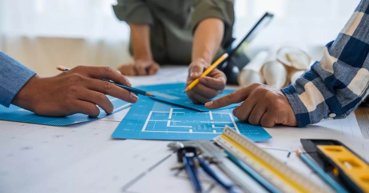 Three people's hands are holding pencils and pointing at a blueprint on a table that includes a level and a compass.