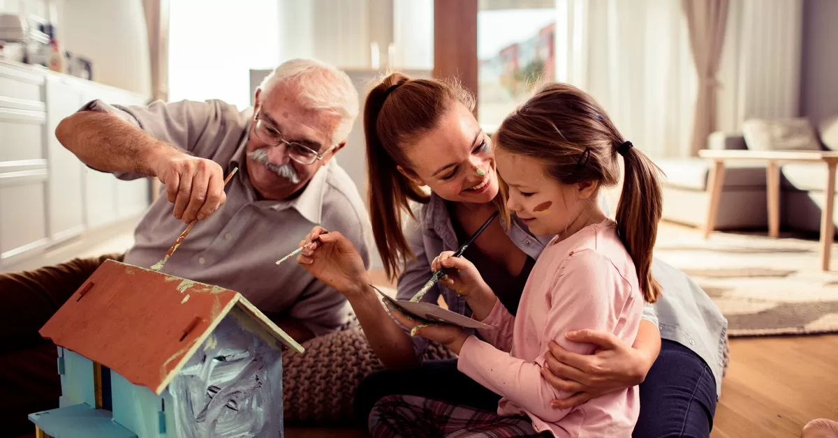 A senior man, a younger woman, and a young girl painting a toy house together while seated on hardwood flooring.