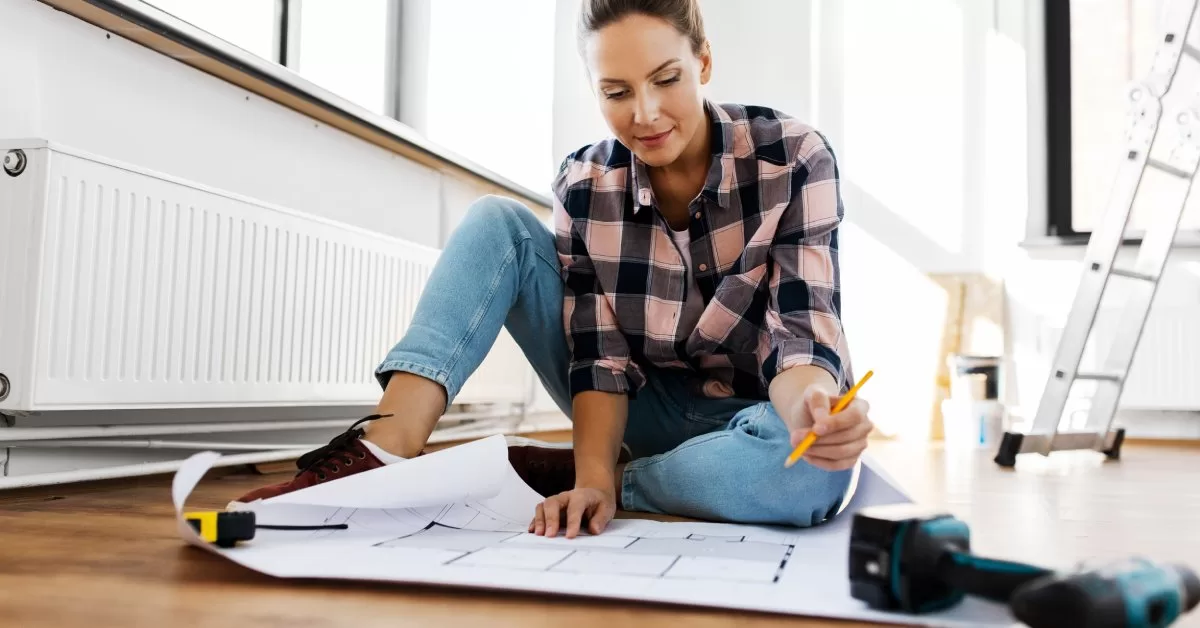 A woman in a pink plaid shirt studies a blueprint unfurled on the floor and weighted with tools. She's holding a pencil.