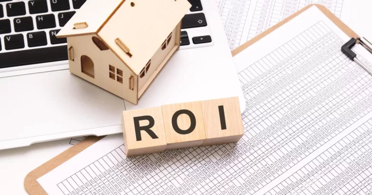 A wooden house figurine rests on a laptop touchpad above printed data tables and wooden cubes spelling “ROI”.