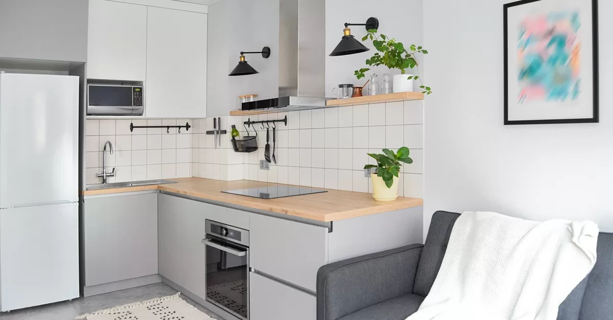 A gray couch sits near white and gray cabinets, a countertop with a stovetop, a microwave, and a range hood.