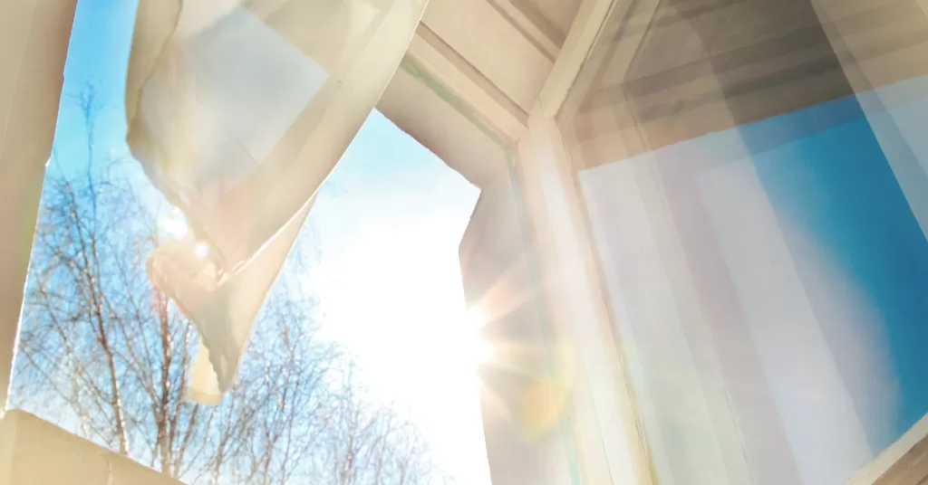 The interior view of a home's open window shows the sun shinning brightly inside. A slight wind causes the curtain to move.