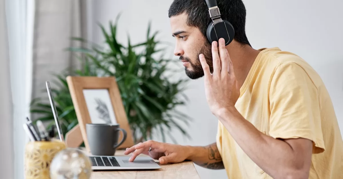 A man wears noise-canceling headphones while working on a laptop at a wooden desk with a plant, pen hold, and photo frame.