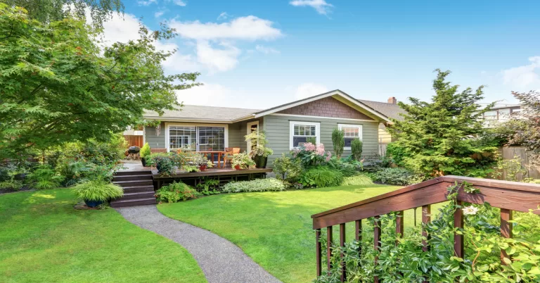 A sage green ADU contains white trim, a wooden deck, potted plants, and a lush garden under a bright blue sky.